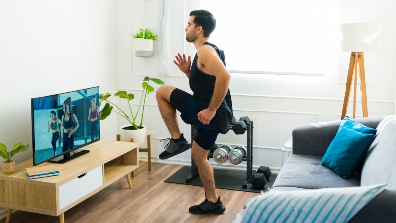 Moški v dnevni sobi izvaja vaje in sledi digitalni vadbi, ki jo gleda na televizijskem zaslonu.