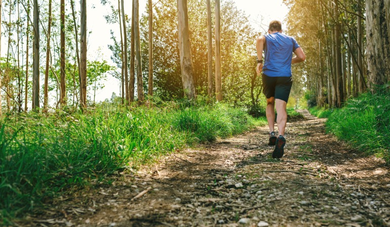 Tekač v modri majici med fartlek treningom teče po razgibani, s soncem obsijani gozdni poti.